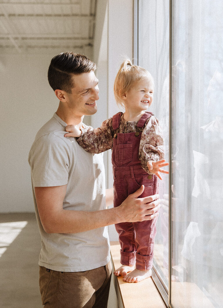 a dad stands near a window with his two year old daughter and they both giggle in the sunshine during a family session by Haverlee Photography