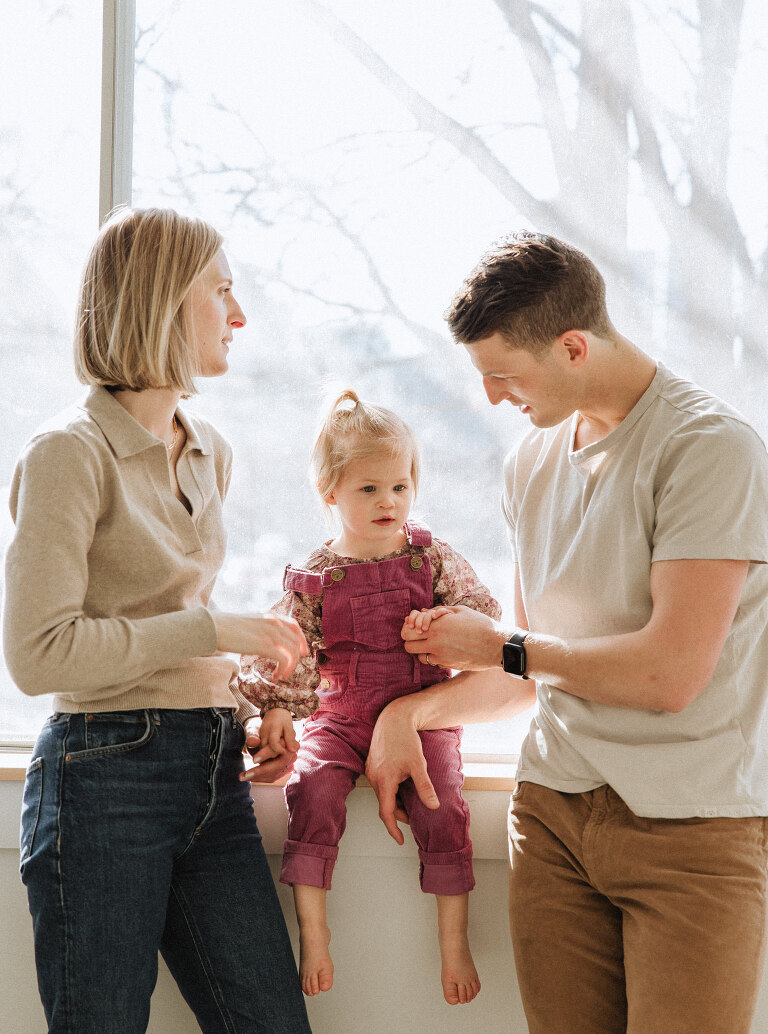 a mom and dad and their two year old daughter in front of a window at East Fifth Studio in downtown Des Moines by wedding and family photographer Haverlee Photography 
