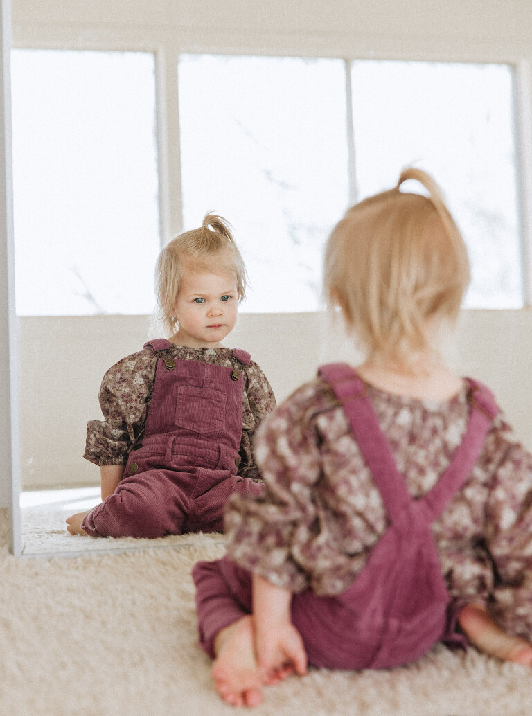 a two year old girl wearing overalls looks in the mirror during a downtown Des Moines family session by Haverlee Photography